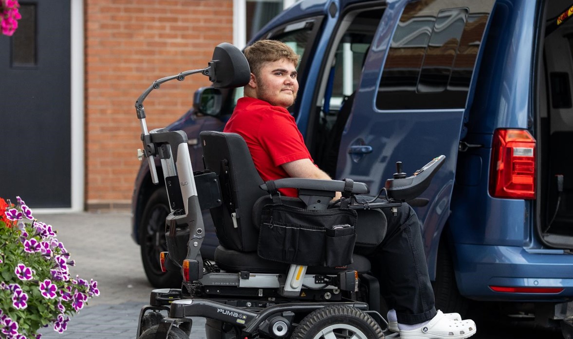 Harry is sitting in his powered wheelchair, outside his house, next to his vehicle. 
