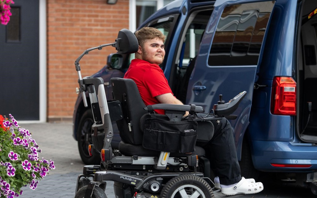 Harry is sitting in his powered wheelchair, outside his house, next to his vehicle. 