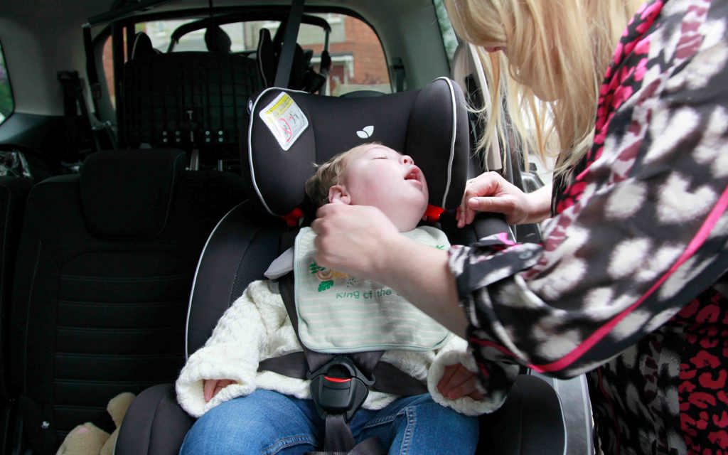 A baby boy is within a car, sitting in a car seat while his mum is securing the car seat safely. 