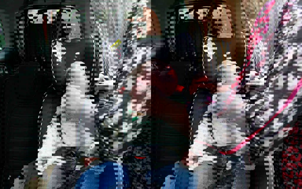 A baby boy is within a car, sitting in a car seat while his mum is securing the car seat safely.