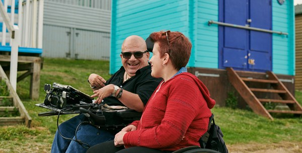 Angela and Pete and both laughing and talking whilst wheeling down the sea front. Behind them are brightly painted beach huts. 