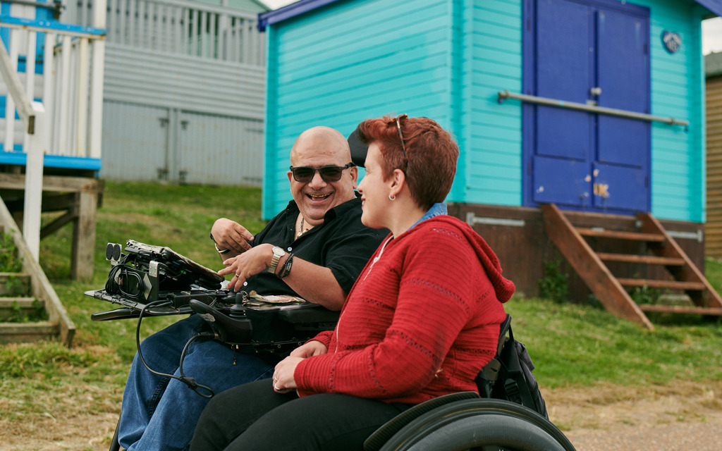 Angela and Pete and both laughing and talking whilst wheeling down the sea front. Behind them are brightly painted beach huts. 