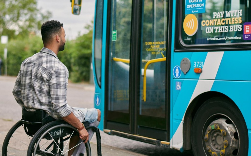 A man with short brown hair is sitting in a wheelchair on the side of the road ready to wheel into a parked blue bus. 