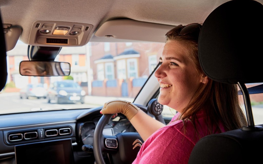 Jess is sitting in her car smiling, looking to her left-hand side. She is wearing a pink dress. 