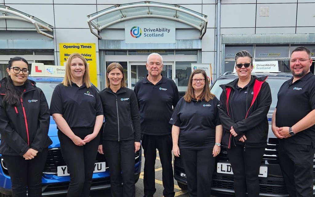 A group photo of five women and two men standing in front of two Driving Mobility Scotland cars smiling in black polos, black trousers and black trainers.