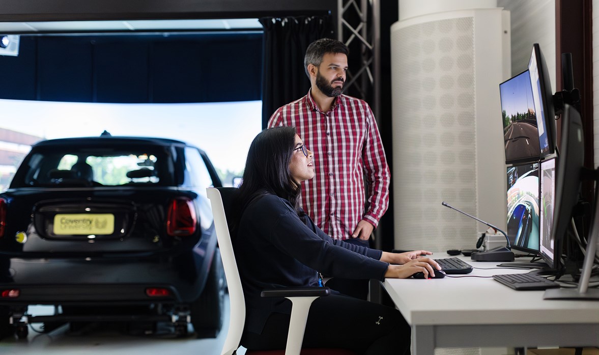 A woman and a man look at a computer screen with a car in the background