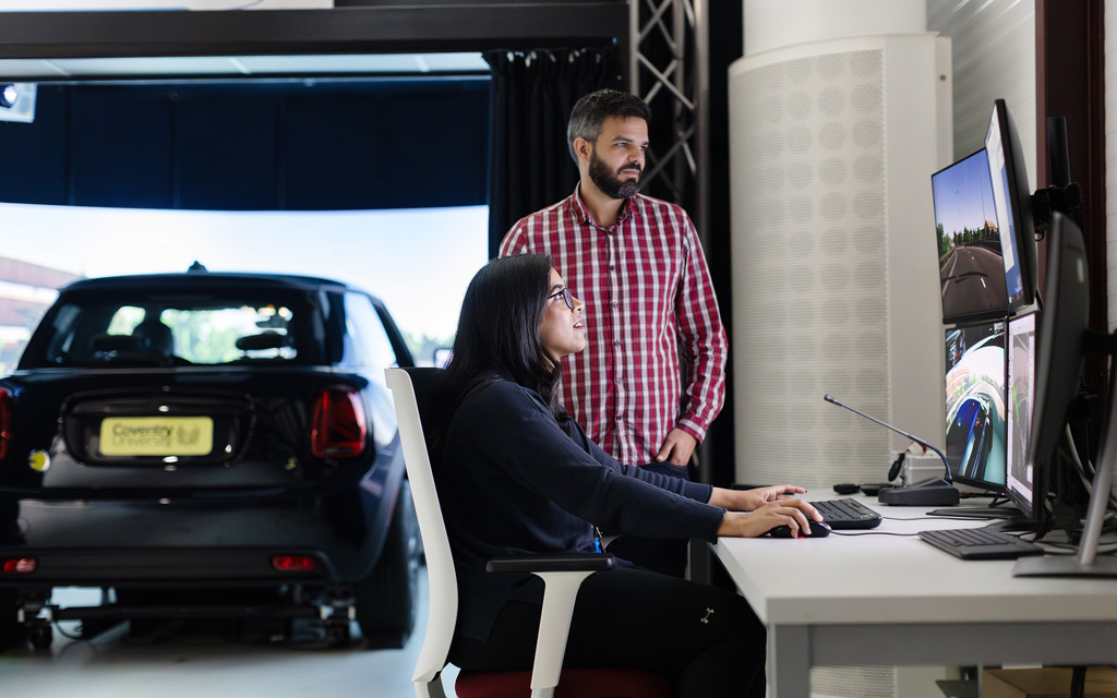 A woman and a man look at a computer screen with a car in the background