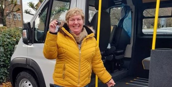 Lady smiling and proudly holding out the keys to a community transport vehicle, which is behind her. 