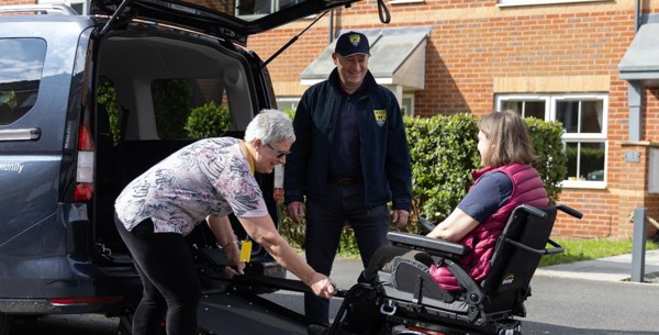 The image shows a gentleman in a navy blue baseball cap, navy blue jacket, black trousers and black trainers standing at the back of a vehicle with the ramp down smiling. An older lady smiling with grey hair, patterned pink top, black trousers and black ballerina pumps is to the right of him attaching a wheelchair to the ramp. In the centre of the image is a women seated in the wheelchair with brown hair, wearing a bright pink gilet.