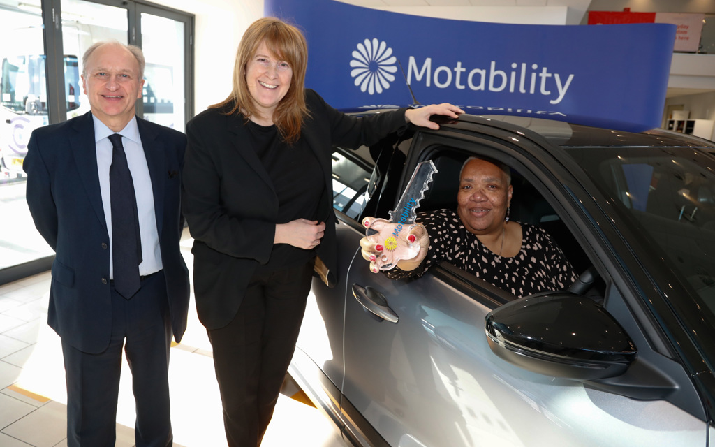 A smiling man and woman stand next to another woman sitting in a shiny new car holding a big key