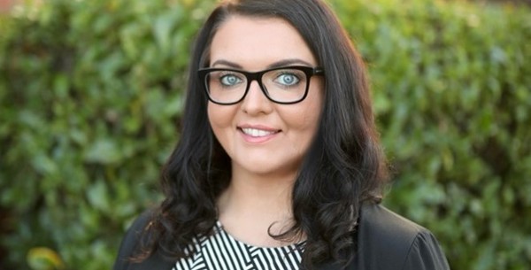 Emma Partlow ncat CEO is smiling in a black blazer and white and black stripped top. She has long brown curly hair and black rimmed glasses.