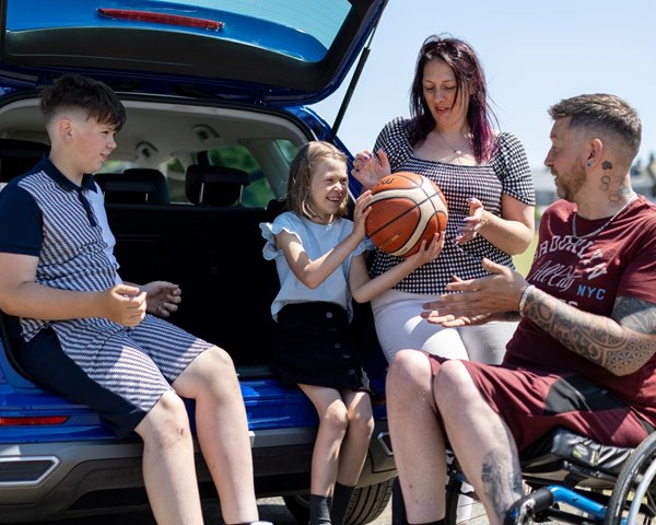 Aaron is sitting in his wheelchair next to his car. The car boot is open and his children are sitting in it. Whilst his wife stands next to the car. They are all talking to one another.